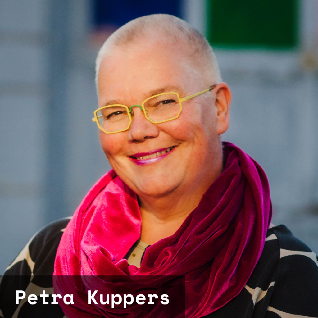 Petra Kuppers, a white queer cis disabled woman of size, head tilted, smiles with twinkling eyes. She has yellow glasses, a shaved head, pink lipstick, purple scarf, polka-dot top, and one of her hands caresses the handlebars of Scootie, her mobility scooter, in front of an urban building with colored glass windows.